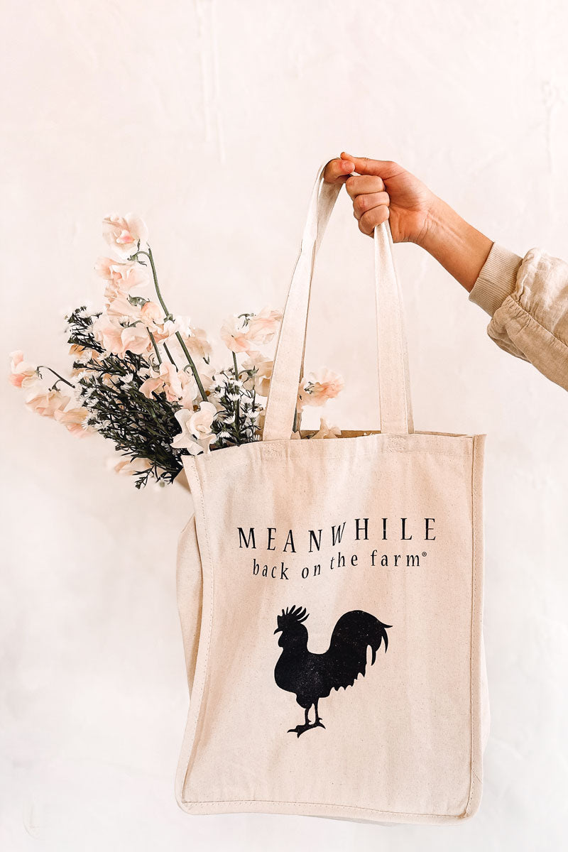 Girl holding a beige Meanwhile back on the farm tote bag with the black rooster logo and with flowers inside