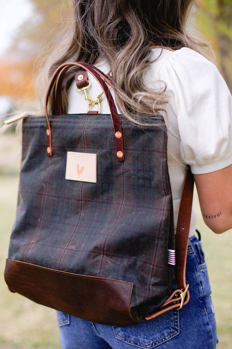 Woman holding a green plaid backpack with meanwhile logo and dark brown straps.