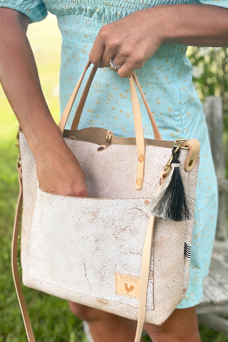 A woman holding the shoulder strap of the birch white bag with a tan body strap, tan tassel, and meanwhile logo. 