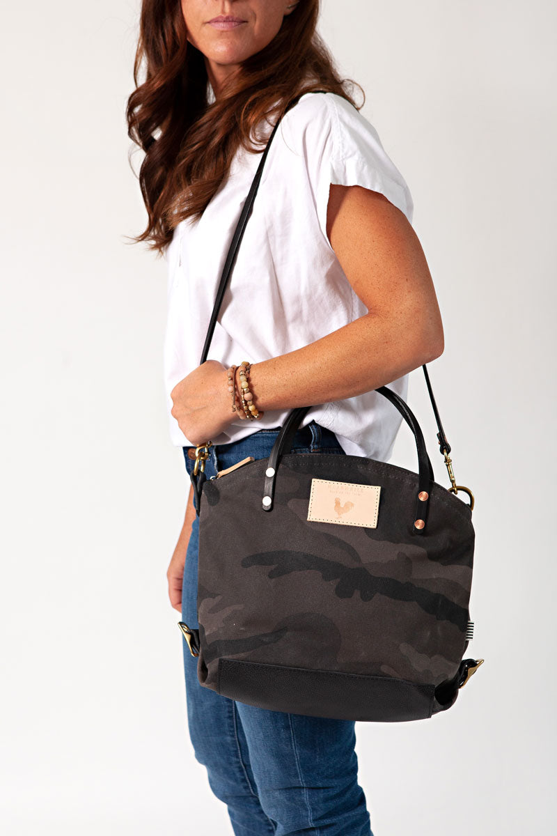 A woman wearing the graphite camouflage backpack with black straps and the meanwhile logo.