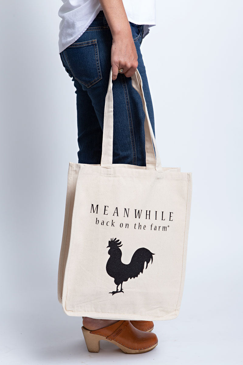 Girl wearing a beige Meanwhile back on the farm tote bag with the black rooster logo with flowers inside
