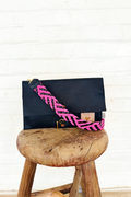 A black leather clutch with a bright pink and black crossbody strap sitting on a stool