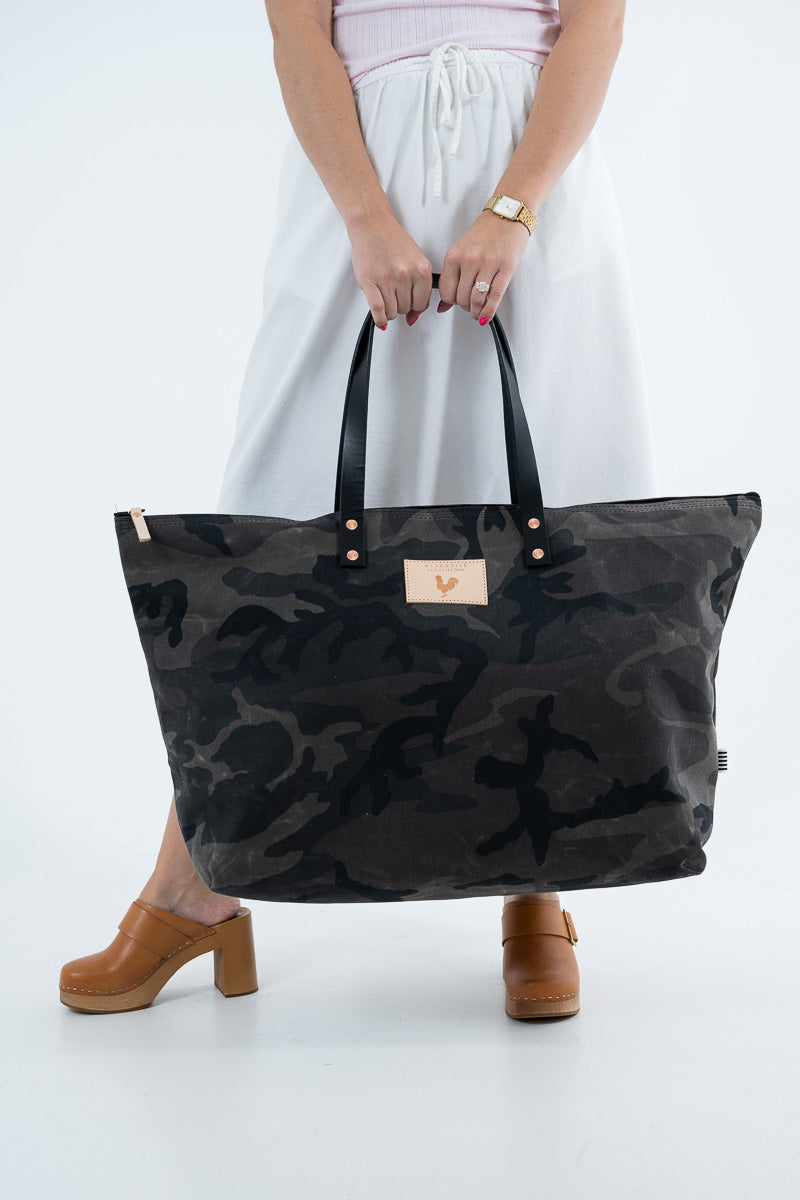 A person holding a large black and dark gray camouflage tote bag with black handles and a small tan logo patch. The person is wearing a white skirt, a light pink top, and brown clogs against a clean white studio background. Handcrafted by Meanwhile Back on the Farm in Lynchburg, VA.