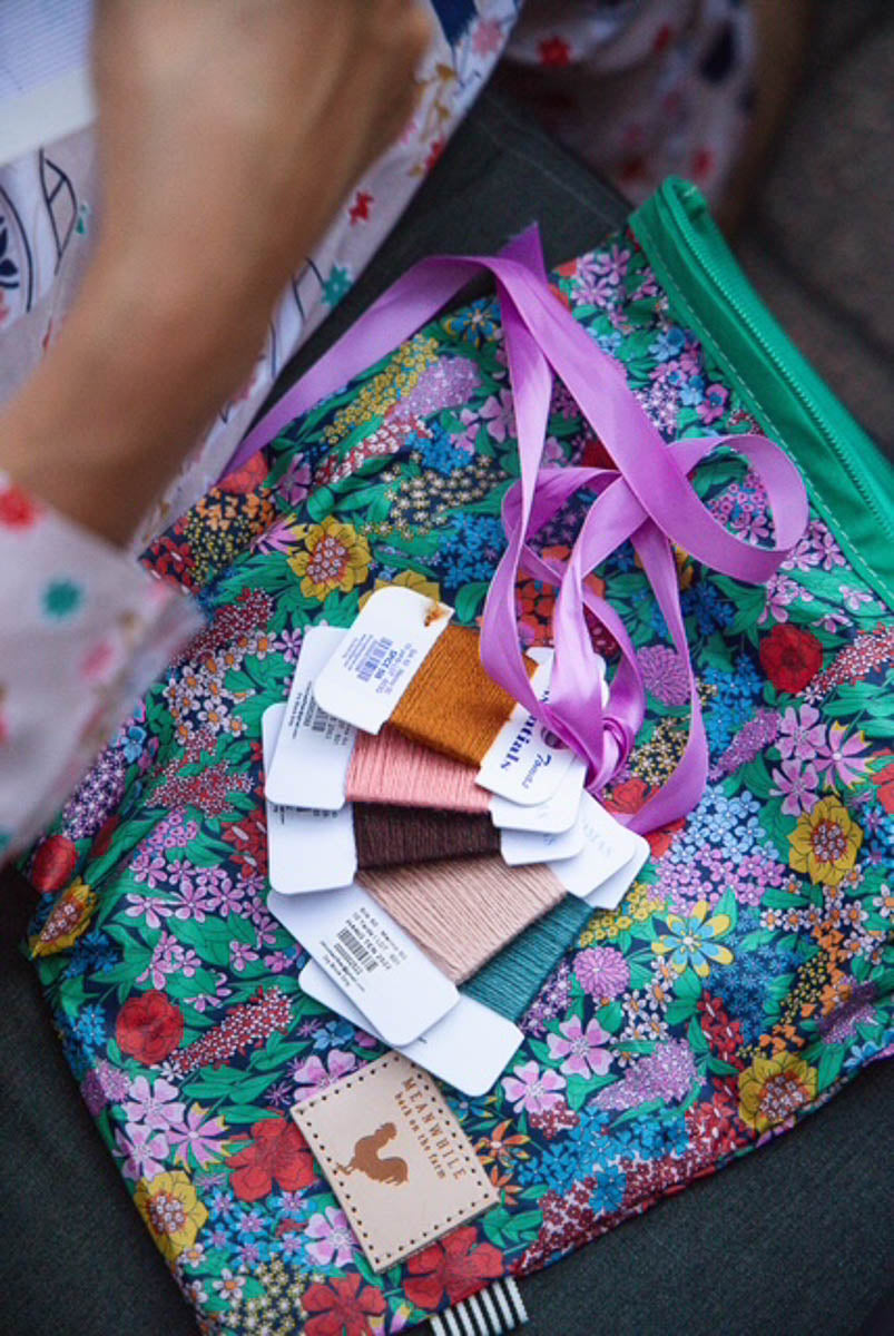 A close-up of a colorful floral zippered pouch with a green zipper, containing several thread samples tied with a purple ribbon. A hand wearing a white floral-patterned sleeve is visible on the left side. Handcrafted by Meanwhile Back on the Farm in Lynchburg, Va.