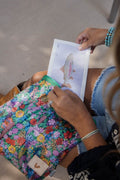 A person holds a printed fish design over a colorful floral fabric pouch or bag. The fabric has a multicolor floral pattern with blue, green, pink, yellow, and red tones, and a small tan brand patch near the bottom. Handcrafted by Meanwhile Back on the Farm in Lynchburg, Va.
