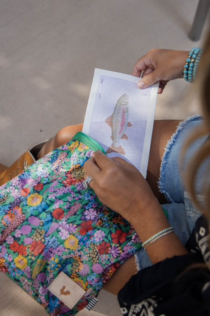 A person holds a printed fish design over a colorful floral fabric pouch or bag. The fabric has a multicolor floral pattern with blue, green, pink, yellow, and red tones, and a small tan brand patch near the bottom. Handcrafted by Meanwhile Back on the Farm in Lynchburg, Va.