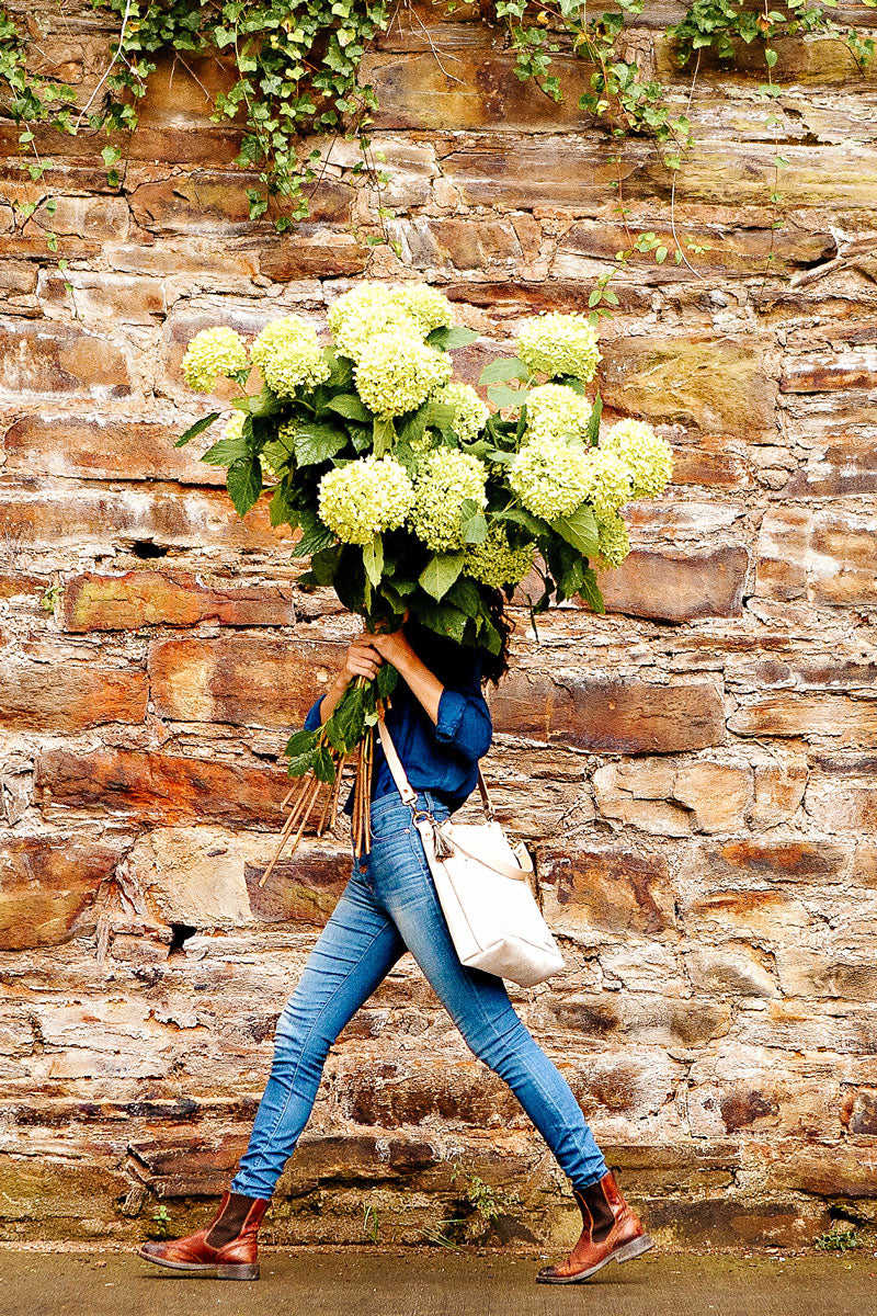 Picture of woman walking briskly in front of a wall carrying a large bundle of white hydrangeas that obscures her face.  She is wearing a large white handbag and cream colored handles and strap.