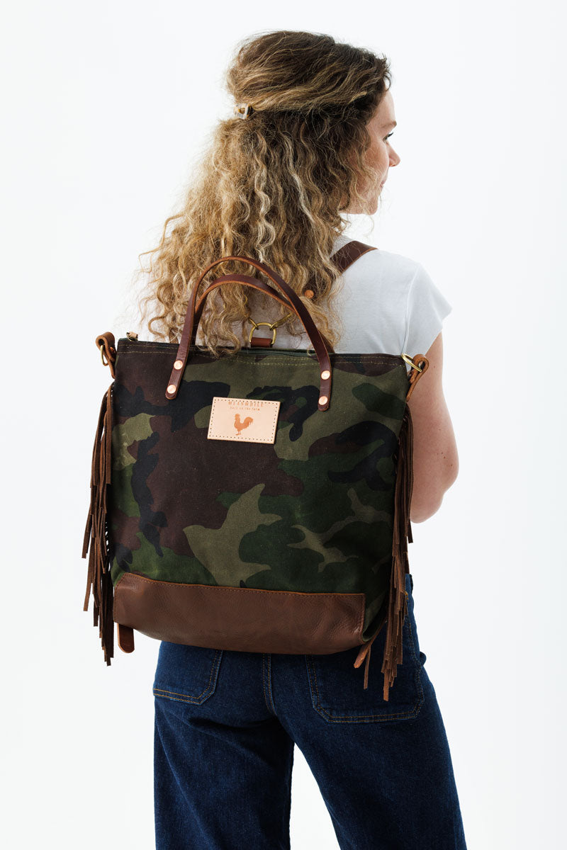 Woman wearing the camouflage bag with the meanwhile logo and brown fringe on her back with backpack straps.