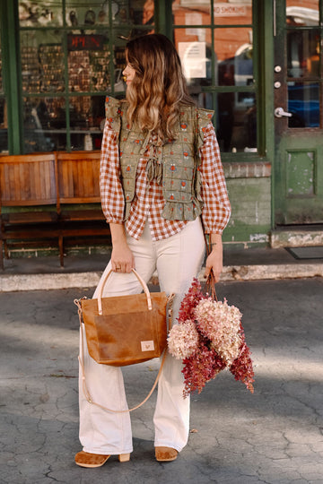 Woman standing in front of a store holding a small brown leather tote and a bouquet of pink/white flowers 