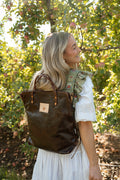 A woman in a white dress stands in an apple orchard wearing a dark brown leather backpack with a small tan square patch on the front. Handcrafted by Meanwhile Back on the Farm in Lynchburg, VA.
