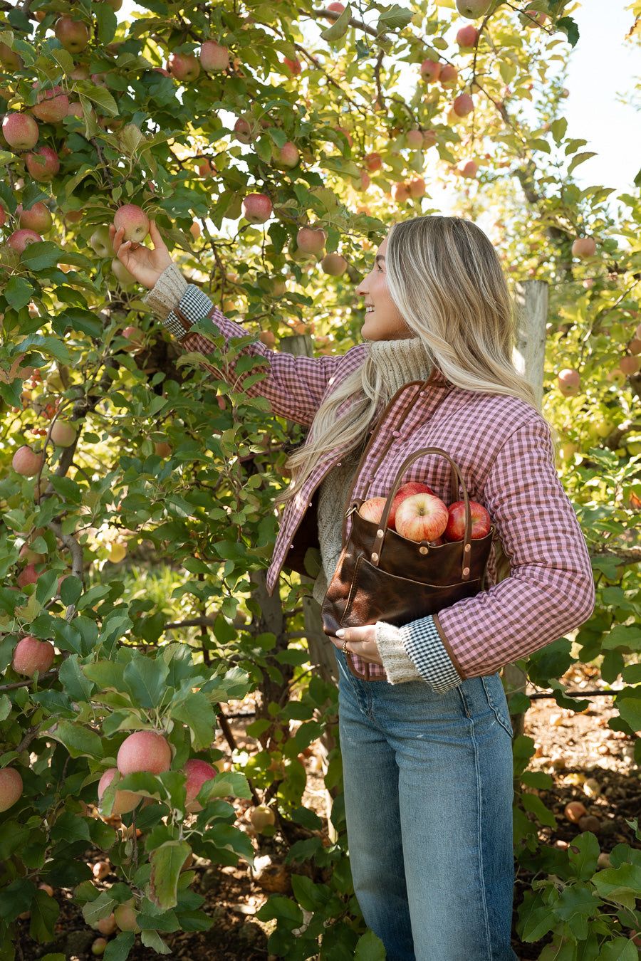 A woman in a red and white gingham shirt and blue jeans stands in an apple orchard, reaching for an apple on a tree while wearing a brown leather crossbody bag filled with apples. Handcrafted by Meanwhile Back on the Farm in Lynchburg, VA.