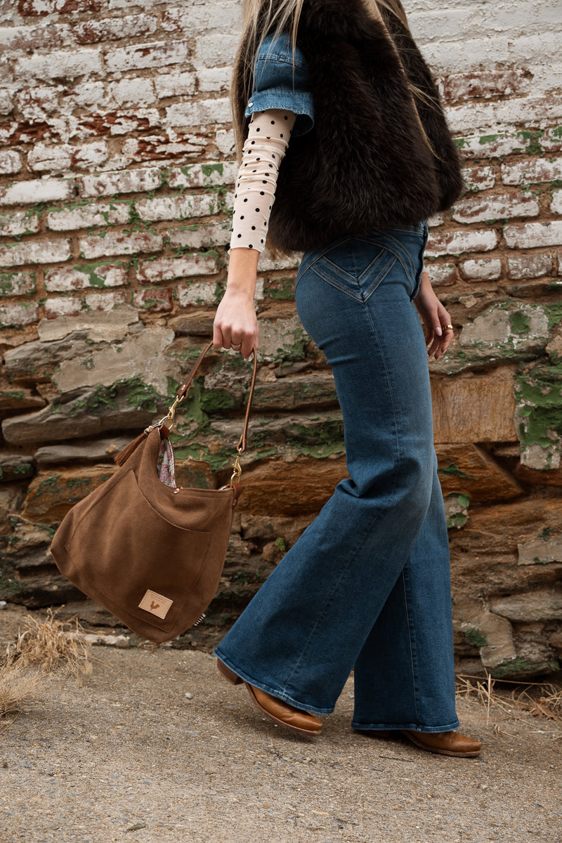 A person wearing blue flare jeans, a dark brown faux-fur vest, and a cream polka-dot long-sleeve shirt walks outdoors against a weathered brick wall while carrying a large brown suede shoulder bag. Handmade by Meanwhile Back on the Farm in Lynchburg,VA