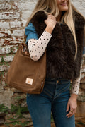 A woman wearing a brown faux fur vest, a blue denim jacket, and a cream polka dot long-sleeve shirt carries a large brown suede shoulder bag with a leather patch on a weathered brick wall background. Handmade by Meanwhile Back on the Farm in Lynchburg,VA
