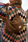 A woman wearing a colorful quilted jacket carries a brown suede crescent shoulder bag with a leather strap and a small brand patch on the front. Handmade by Meanwhile Back on the Farm in Lynchburg,VA