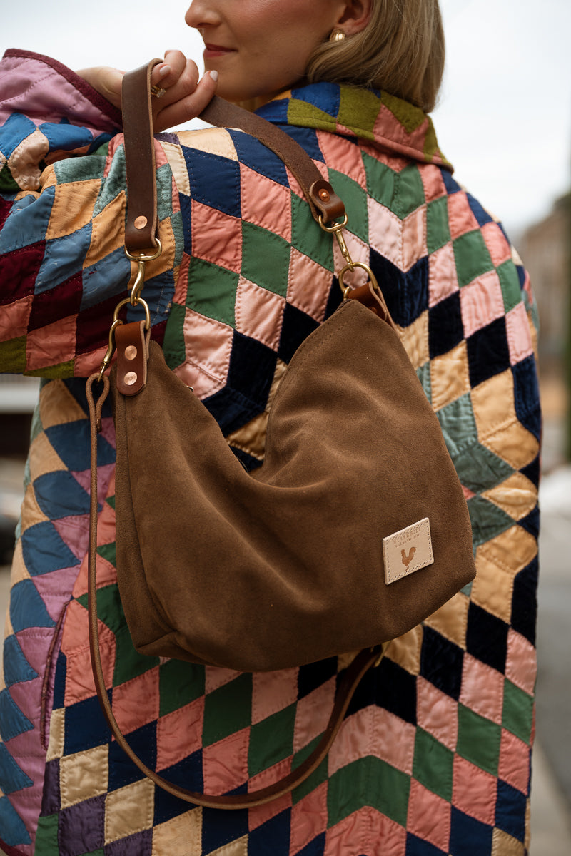 A woman wearing a colorful quilted jacket carries a brown suede crescent shoulder bag with a leather strap and a small brand patch on the front. Handmade by Meanwhile Back on the Farm in Lynchburg,VA