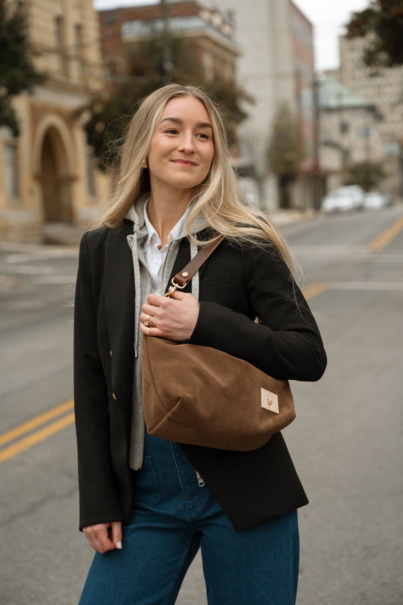 A woman with blonde hair stands on a city street wearing a black blazer, white shirt, blue jeans, and carrying a brown canvas shoulder bag. Handmade by Meanwhile Back on the Farm in Lynchburg,VA
