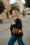 A woman walking on a city street carrying a brown suede shoulder bag with a small white brand patch. She is wearing a black blazer and blue jeans. Handmade by Meanwhile Back on the Farm in Lynchburg,VA