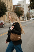 A woman walking on a city street wearing a black blazer, blue jeans, and carrying a brown canvas shoulder bag with a small white logo patch. Handmade by Meanwhile Back on the Farm in Lynchburg,VA