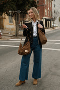 A woman stands on a city street wearing a black double-breasted blazer, white button-down shirt, blue wide-leg jeans, and tan ankle boots, while carrying two brown suede shoulder bags. Handmade by Meanwhile Back on the Farm in Lynchburg,VA