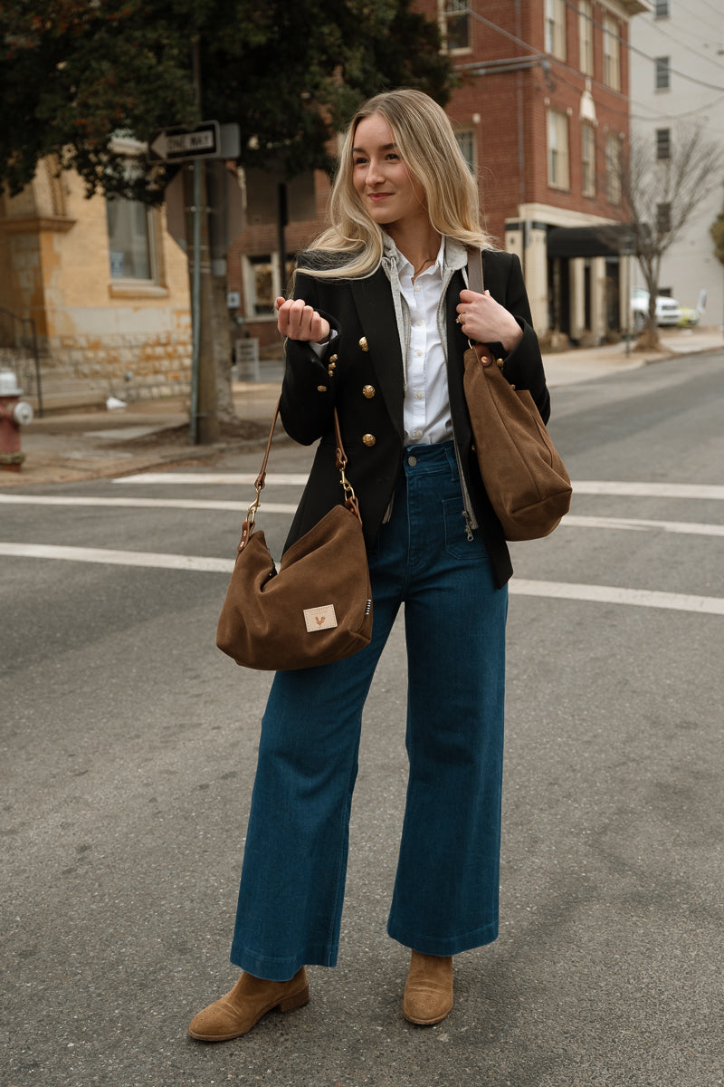 A woman stands on a city street wearing a black double-breasted blazer, white button-down shirt, blue wide-leg jeans, and tan ankle boots, while carrying two brown suede shoulder bags. Handmade by Meanwhile Back on the Farm in Lynchburg,VA