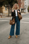 A woman stands on an urban street crosswalk wearing a black blazer, white button-down shirt, wide-leg blue jeans, and tan boots, while carrying two brown suede shoulder bags. Handmade by Meanwhile Back on the Farm in Lynchburg,VA