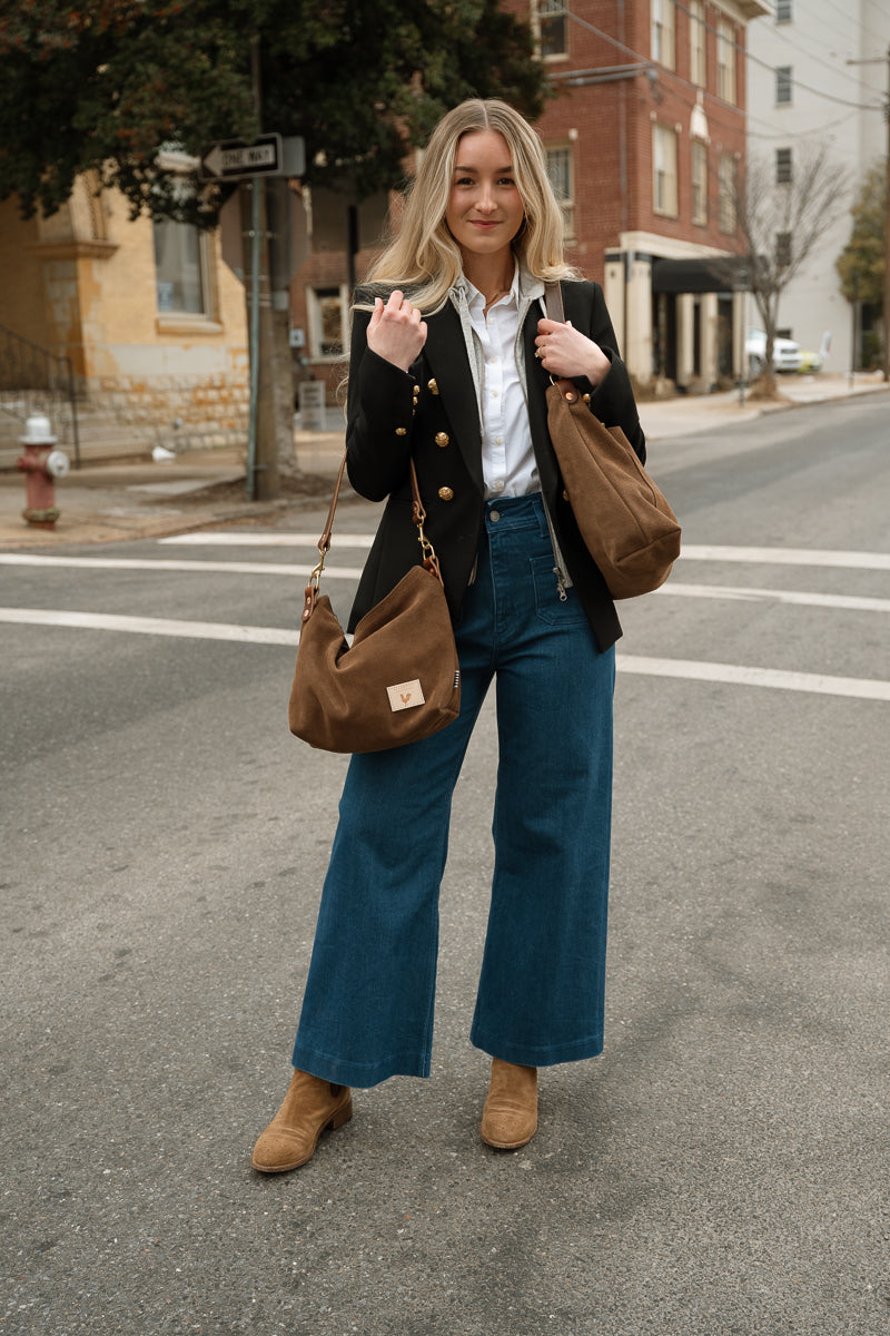 A woman stands on an urban street crosswalk wearing a black blazer, white button-down shirt, wide-leg blue jeans, and tan boots, while carrying two brown suede shoulder bags. Handmade by Meanwhile Back on the Farm in Lynchburg,VA