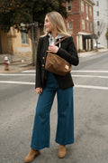 A woman stands on a city street wearing a black blazer with gold buttons, a white shirt, blue wide-leg jeans, and tan boots, while carrying a brown canvas shoulder bagHandmade by Meanwhile Back on the Farm in Lynchburg,VA