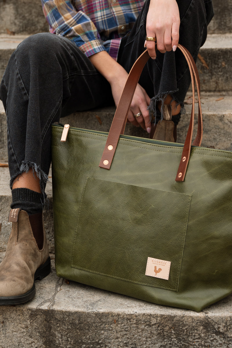 An olive green canvas tote bag with brown leather handles and a small tan brand patch, shown being held by a person sitting on stone steps. Handcrafted by Meanwhile Back on the Farm in Lynchburg, VA.