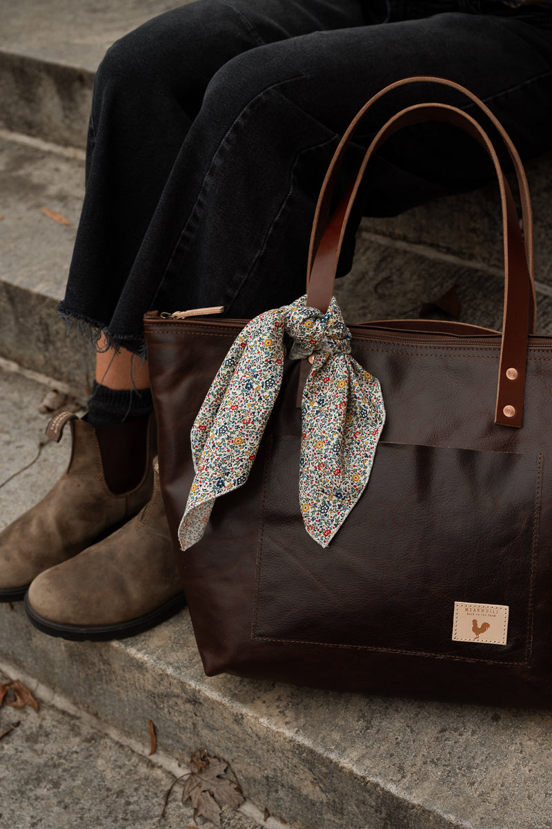A woman wearing a colorful plaid shirt and black jeans carries a large dark brown leather tote bag with brown shoulder straps and metal rivets, standing on outdoor stone steps. Handcrafted by Meanwhile Back on the Farm in Lynchburg, VA.