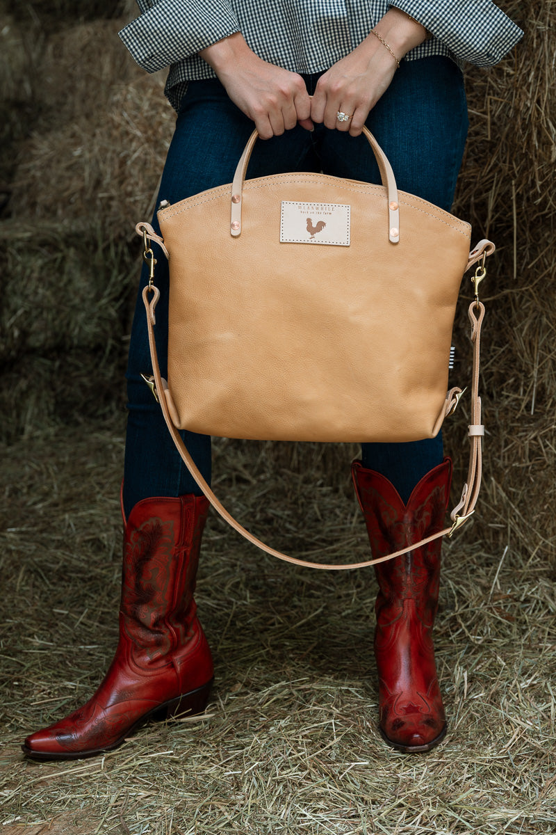 A woman wearing a white ruffled top and blue jeans carries a tan leather backpack with top handles and shoulder straps against a plain white background. Handmade by Meanwhile Back on the Farm in Lynchburg,VA