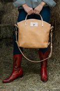 A woman standing in front of hay bales holding a tan leather bag and wearing red leather cowgirl boots. Handcrafted by Meanwhile Back on the Farm in Lynchburg, VA.