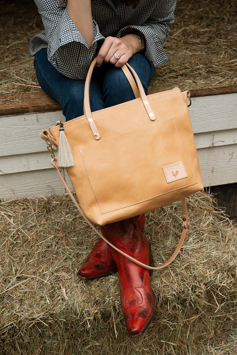 A woman sitting on a ledge in hay holding a tan leather bag and wearing bright red cowgirl boots.