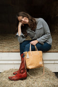 A woman sitting casually in a barn holding a tan leather bag and wearing a button up shirt, blue jeans and red cowgirl boots. Handcrafted by Meanwhile Back on the Farm in Lynchburg, VA.