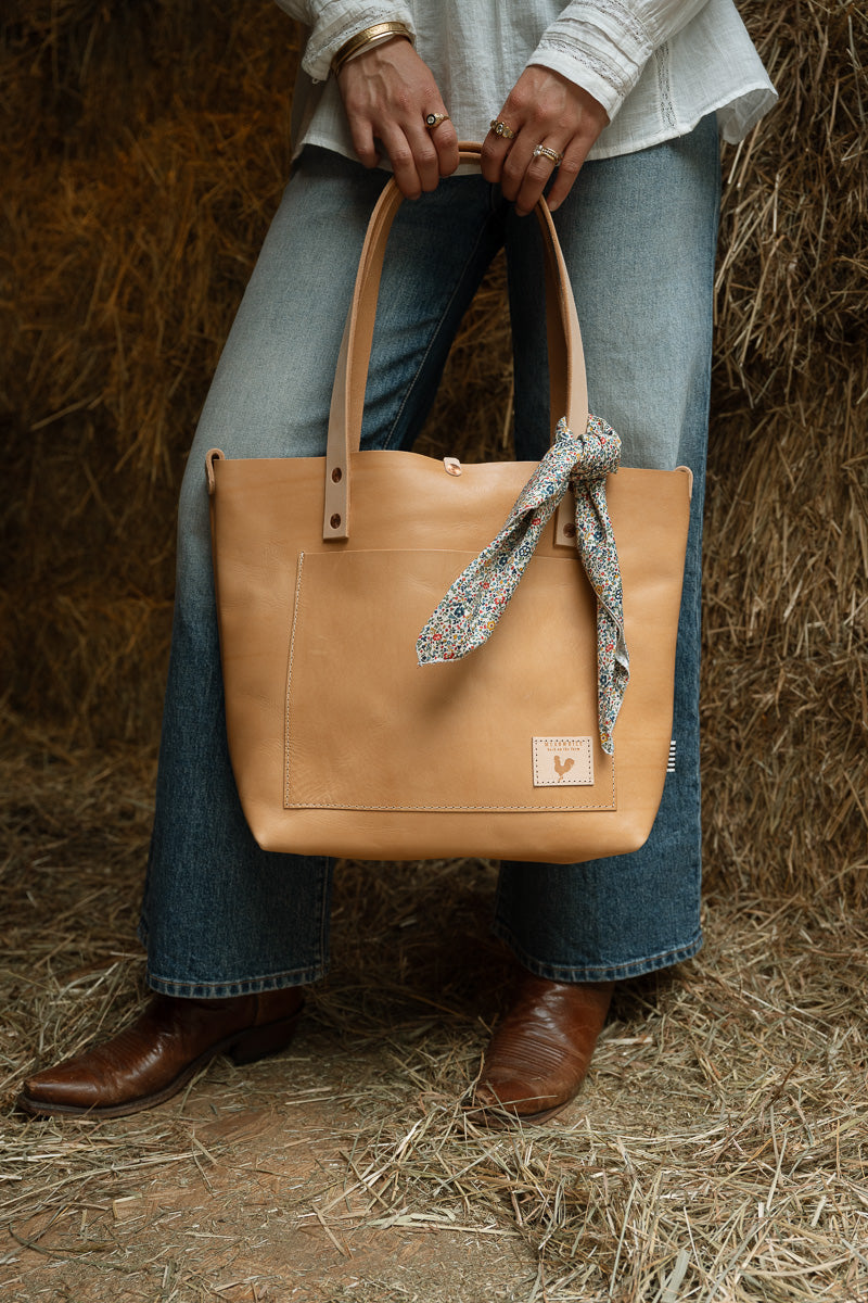 A woman in front of hay bales holding a tan leather bag with a floral scarf tied to the handle. Handcrafted by Meanwhile Back on the Farm in Lynchburg, VA.