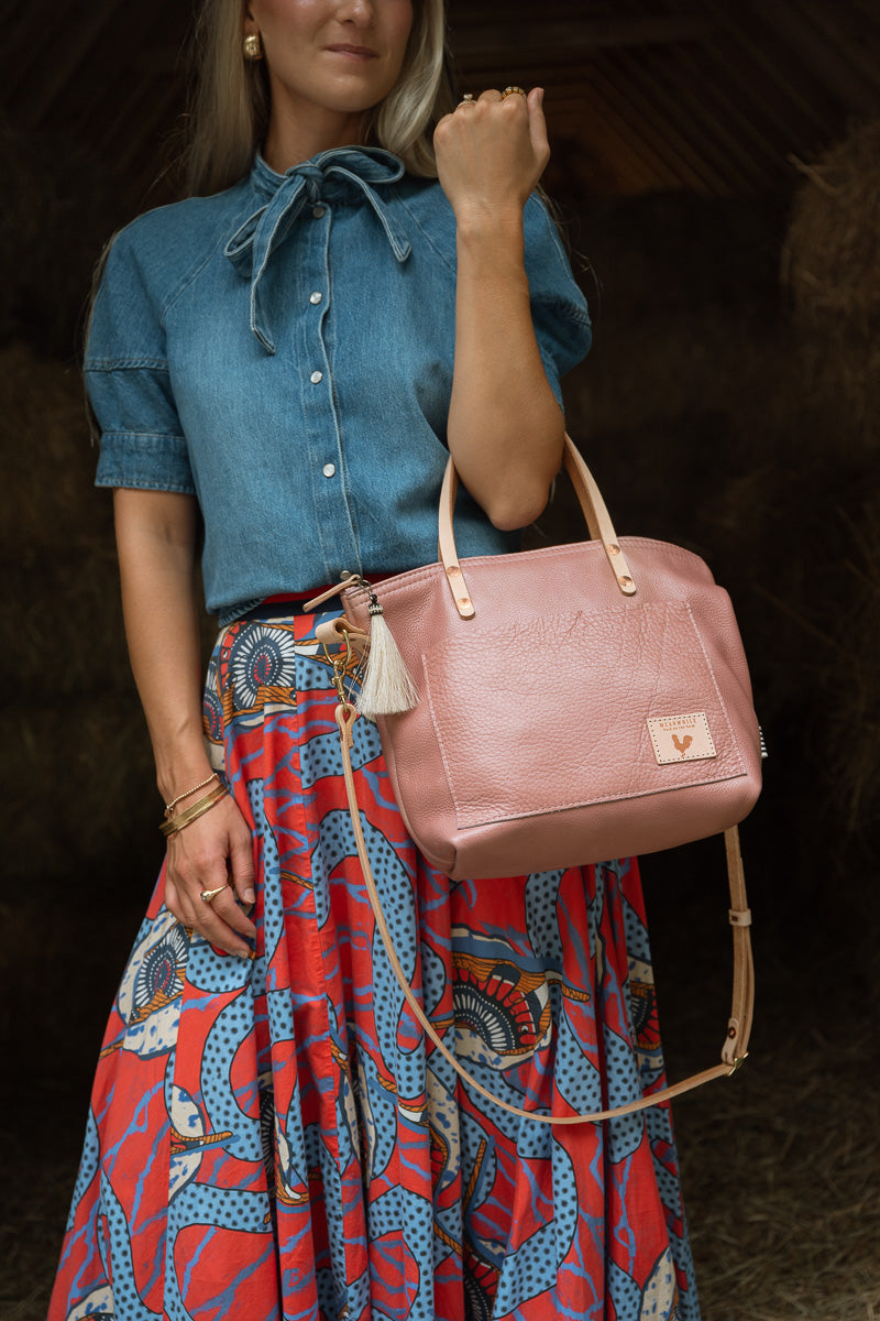 A woman in a blue button up shirt and long red skirt modeling a pink leather handbag with tan leather straps. Handcrafted by Meanwhile Back on the Farm in Lynchburg, VA.