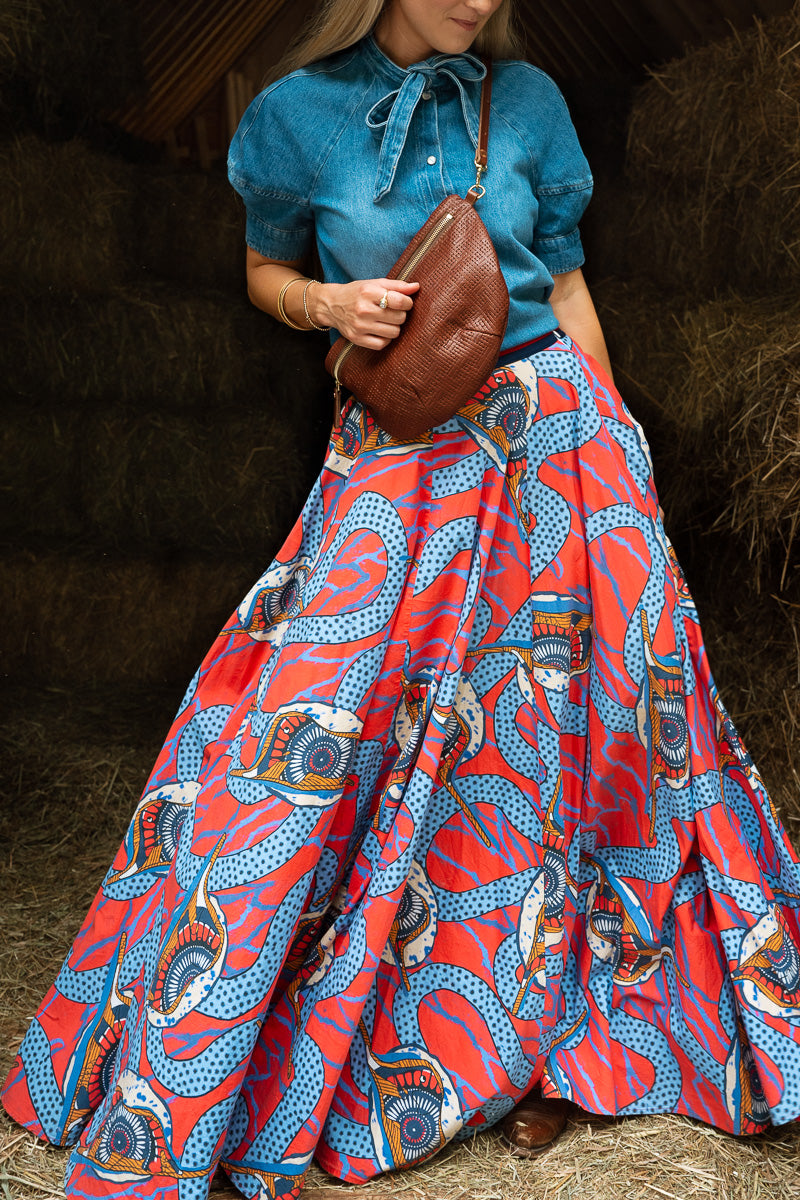 A woman wearing a vibrant blue and red patterned African wax print maxi skirt with a matching blue tie-neck blouse, holding a brown leather shoulder bag in a rustic barn setting. Handcrafted by Meanwhile Back on the Farm in Lynchburg, VA.