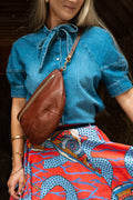 A woman wearing a blue denim shirt and a vibrant red, blue, and yellow patterned skirt, carrying a brown textured crocodile-embossed crossbody bag. Handcrafted by Meanwhile Back on the Farm in Lynchburg, VA.