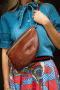 A brown textured leather crossbody bag with gold-tone hardware, worn across the waist of a model wearing a blue denim shirt and a colorful patterned skirt. Handcrafted by Meanwhile Back on the Farm in Lynchburg, VA.