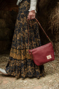 A woman in a long patterned skirt holding a small red leather handbag and a long red leather strap. Handcrafted by Meanwhile Back on the Farm in Lynchburg, VA.