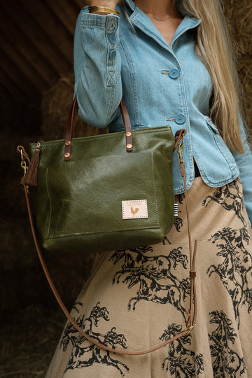 A woman in a button up blue shirt and patterned skirt modeling an olive green leather bag with dark brown leather straps. Handcrafted by Meanwhile Back on the Farm in Lynchburg, VA.