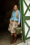 A woman stands in a barn doorway wearing a light blue denim jacket, a beige floral print skirt, and brown cowboy boots, while holding an olive green leather tote bag. Handcrafted by Meanwhile Back on the Farm in Lynchburg, VA.