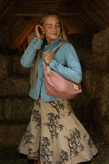 A woman in a blue button up shirt and a tan skirt modeling a pink leather bag with tan leather straps. Handcrafted by Meanwhile Back on the Farm in Lynchburg, VA.