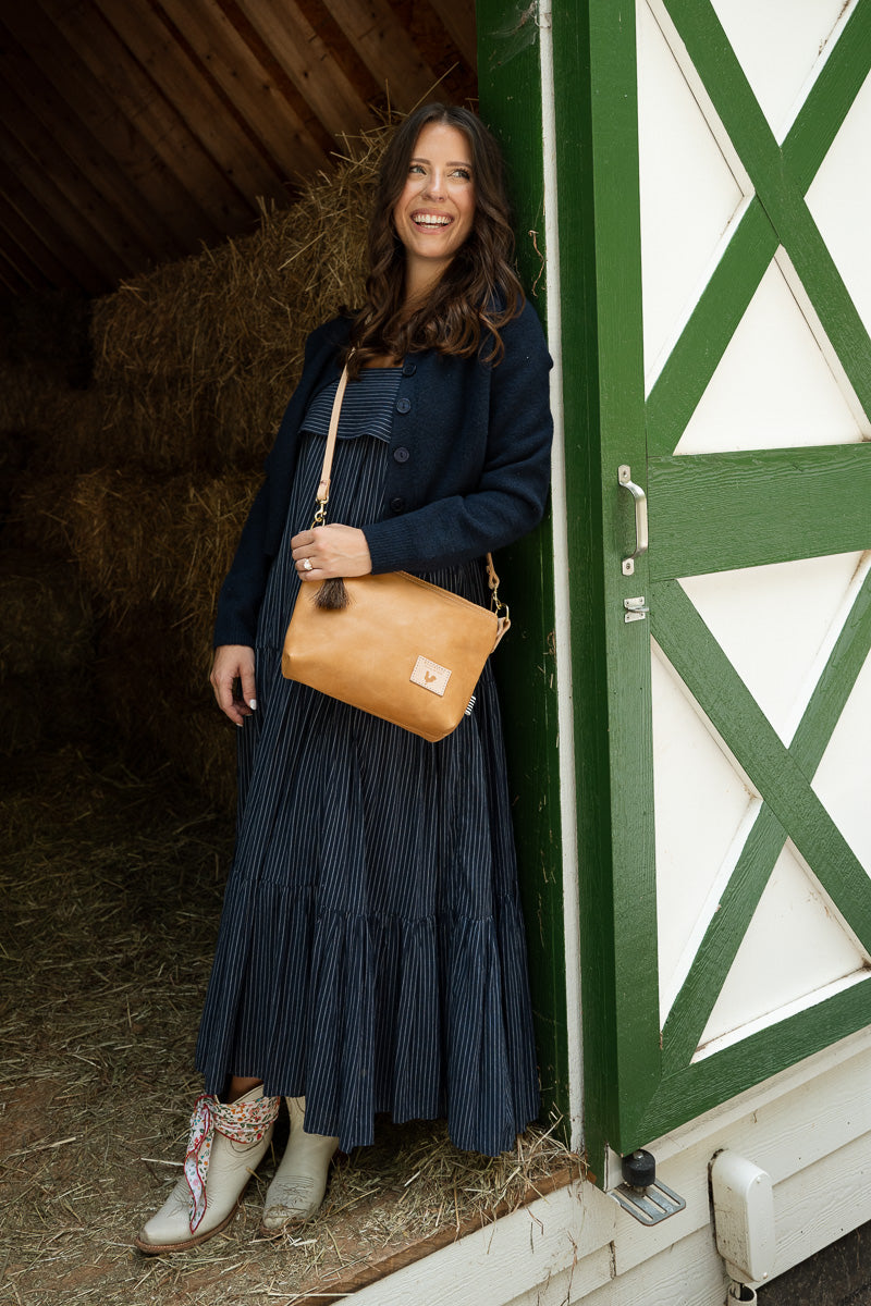 A woman stands in a barn doorway wearing a navy blue pleated midi dress, a navy cardigan, and light brown leather cowboy boots, carrying a tan leather shoulder bag. The background features a green and white barn door and hay bales. Handcrafted by Meanwhile Back on the Farm in Lynchburg, VA.