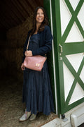 A woman in a blue dress looking out a barn door and modeling a pink leather purse with tan leather straps. Handcrafted by Meanwhile Back on the Farm in Lynchburg, VA.