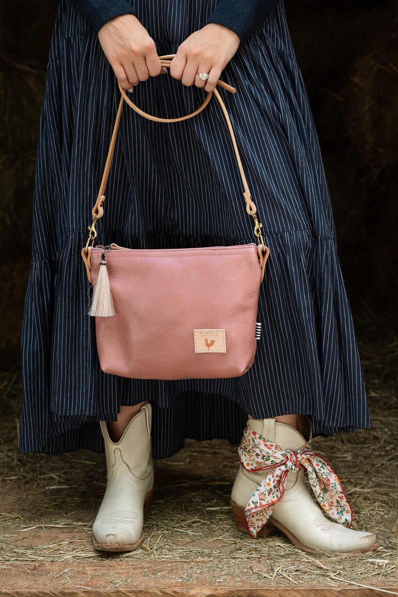 A woman in a blue dress and white cowgirl boots holding a small pink handbag with tan leather straps. Handcrafted by Meanwhile Back on the Farm in Lynchburg, VA.