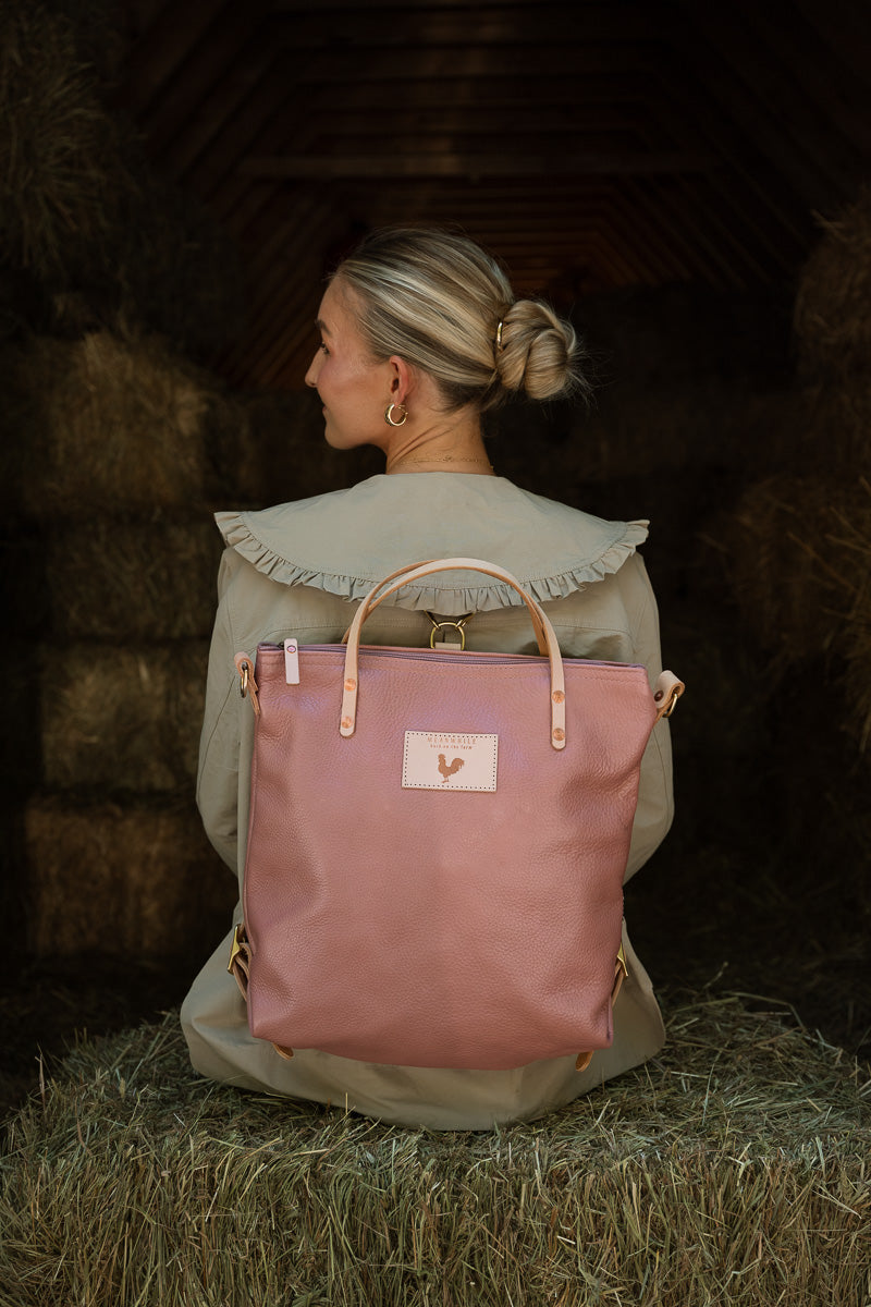A woman with a bun in a tan frilled shirt sitting on a hay bale with her back to the camera. She is wearing a pink leather backpack with tan straps.
