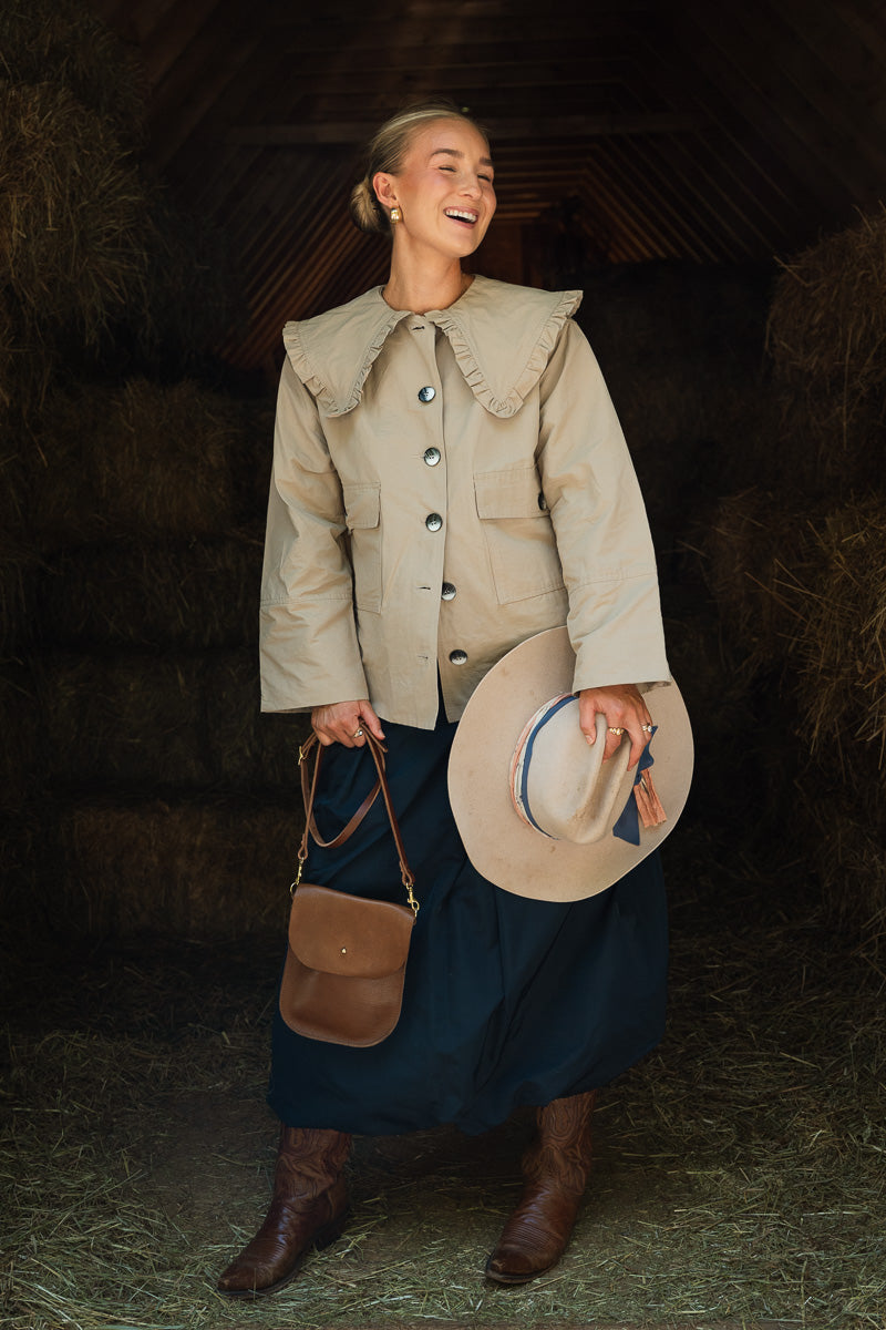 A woman stands in a rustic barn holding a wide-brim hat and a small brown crossbody bag. She is wearing a beige ruffled-collar jacket, a dark navy skirt, and brown boots. Handcrafted by Meanwhile Back on the Farm in Lynchburg, VA.
