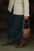 A person wearing a beige jacket, dark navy skirt, and brown leather cowboy boots holds a brown leather shoulder bag in a rustic barn setting. Handcrafted by Meanwhile Back on the Farm in Lynchburg, VA.