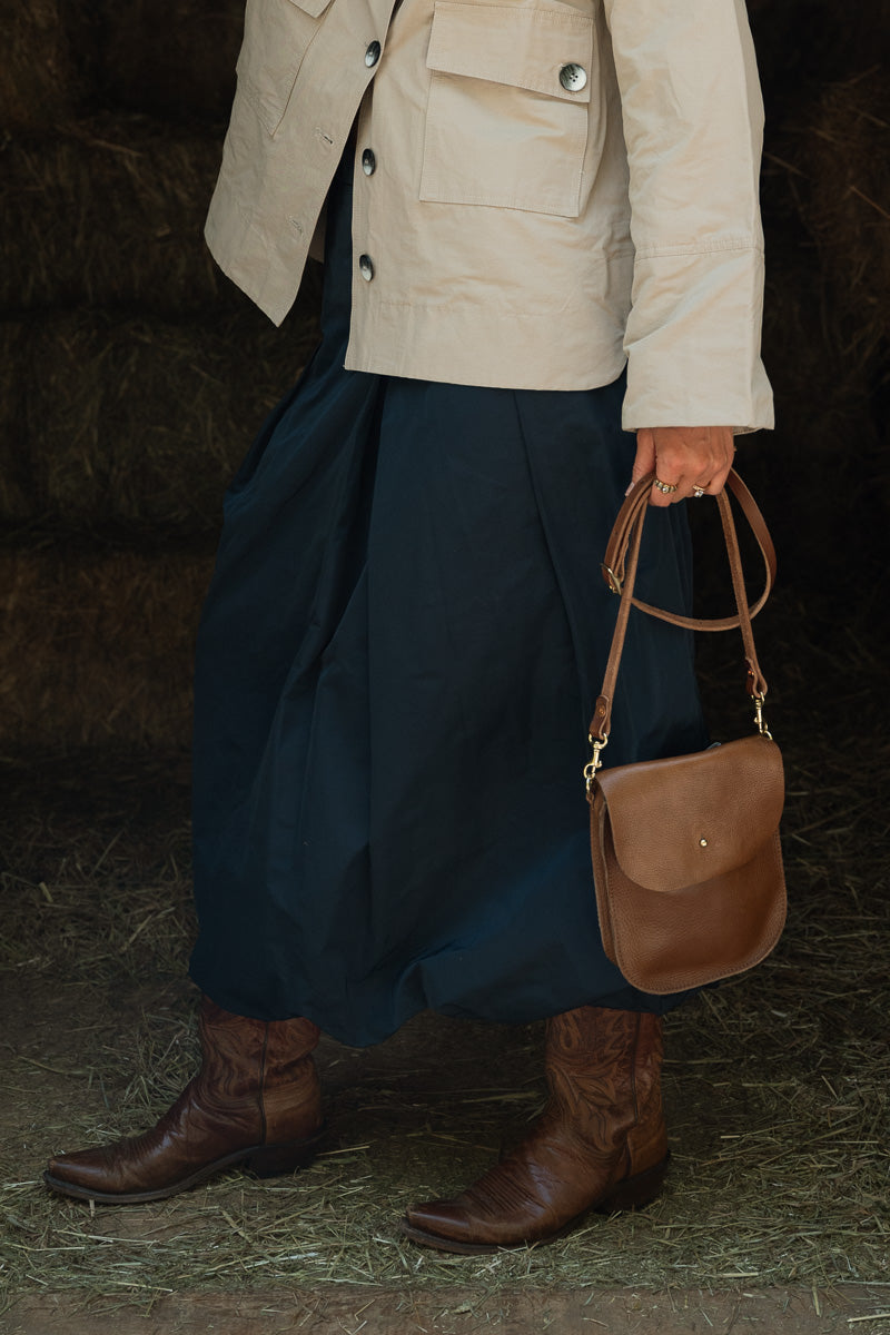A person wearing a beige jacket, dark navy skirt, and brown leather cowboy boots holds a brown leather shoulder bag in a rustic barn setting. Handcrafted by Meanwhile Back on the Farm in Lynchburg, VA.