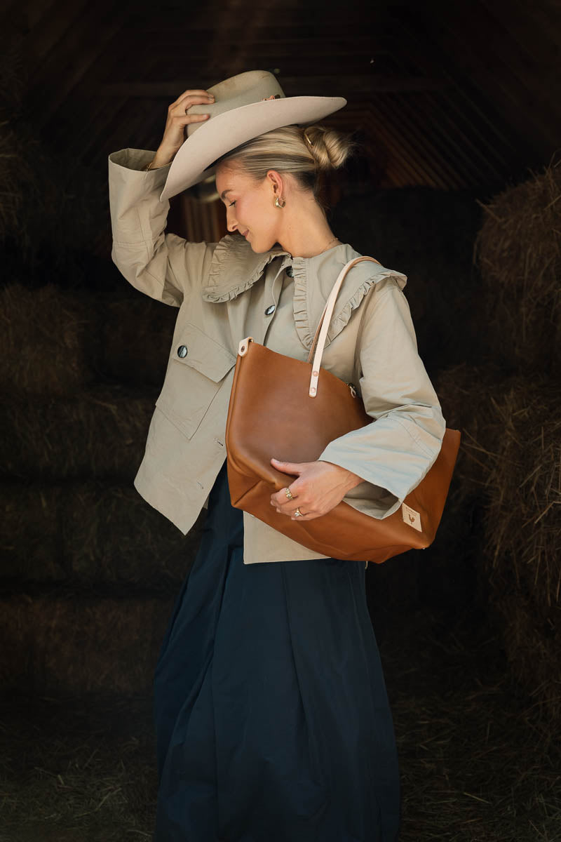 A woman in a rustic barn setting wearing a beige wide-brim hat, a light beige ruffled blouse, a beige double-breasted jacket, and a dark navy skirt, carrying a large brown leather shoulder bag. Handcrafted by Meanwhile Back on the Farm in Lynchburg, VA.
